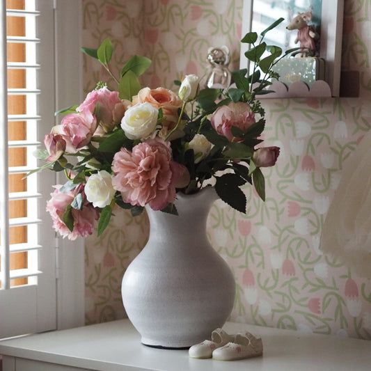 White vase with pink and white flowers on a surface with patterned wallpaper in the background